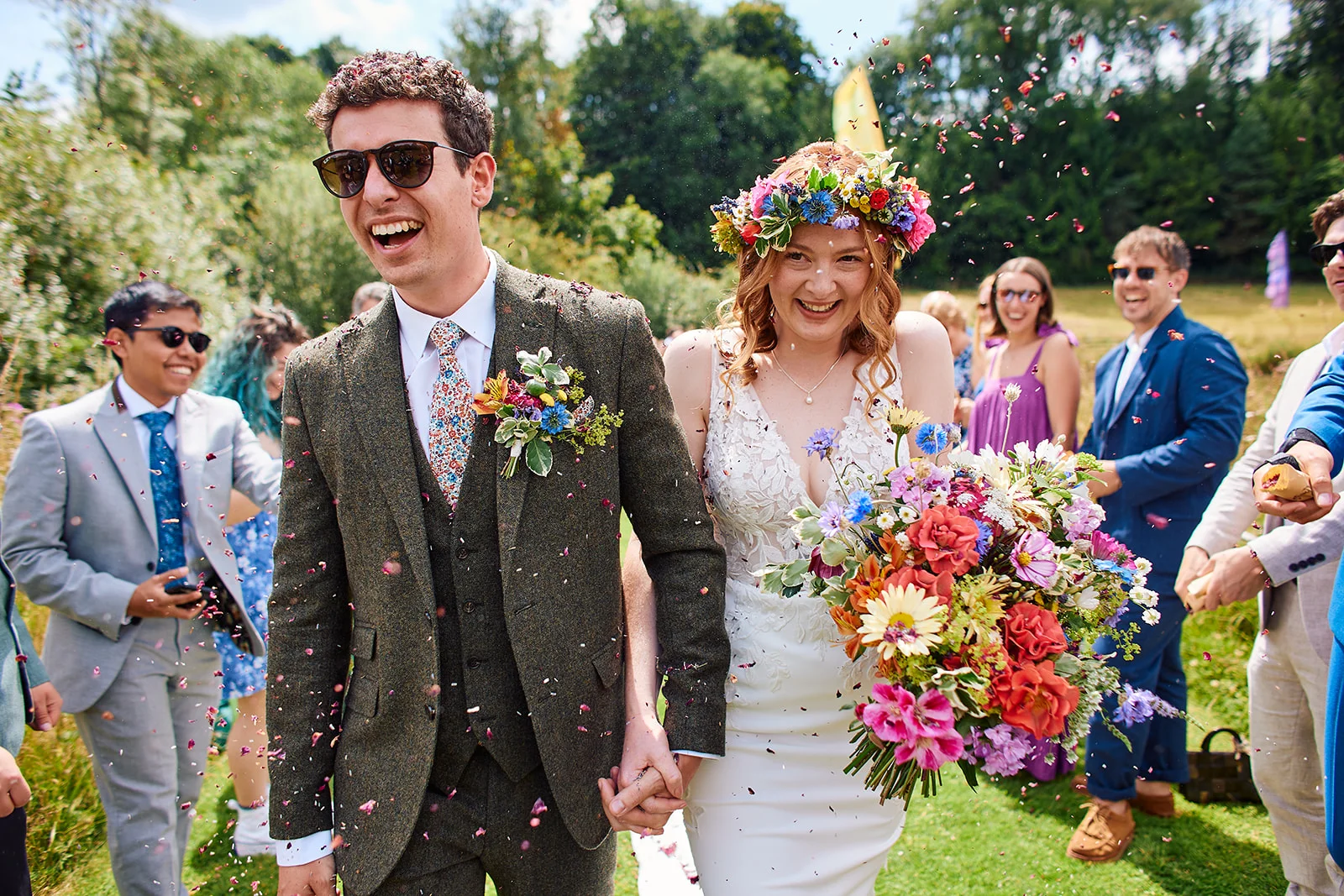 A joyous newlywed couple walk hand in hand through a shower of colourful confetti, smiling widely as wedding guests cheer in a sunlit outdoor garden setting.