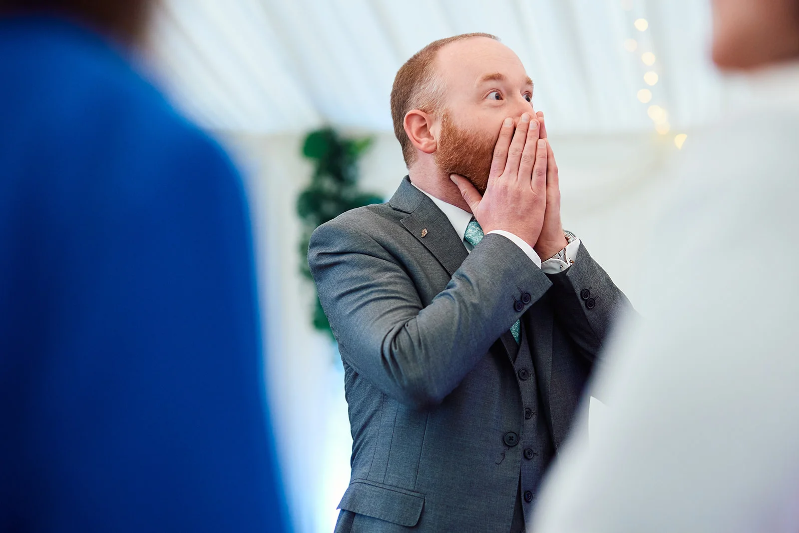 A groom in a grey three-piece suit reacts with sheer surprise and delight, covering his mouth with his hands during the wedding ceremony.