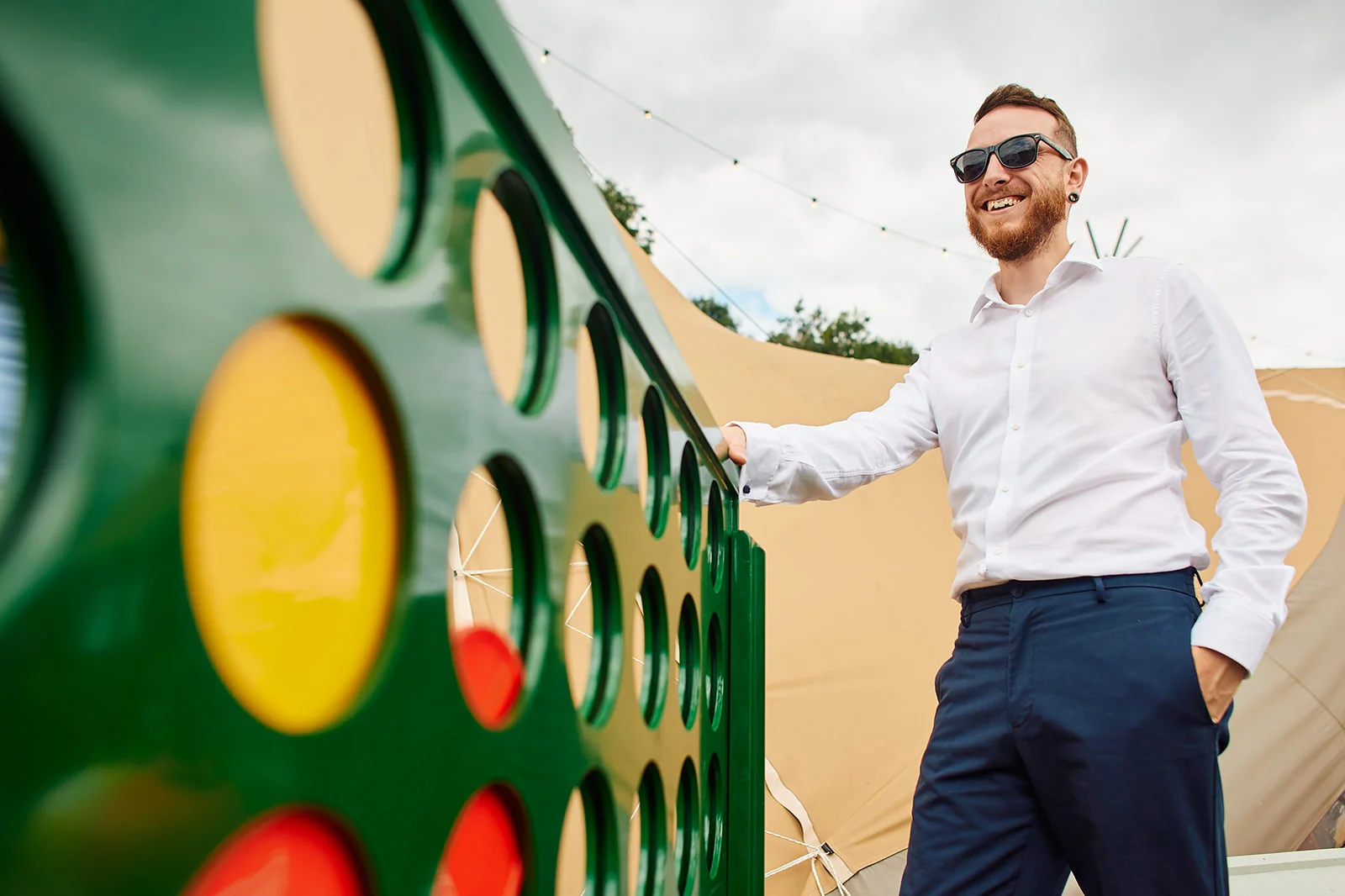 A smiling groom wearing sunglasses and a white shirt stands beside a giant four-in-a-row lawn game outside a wedding tipi.