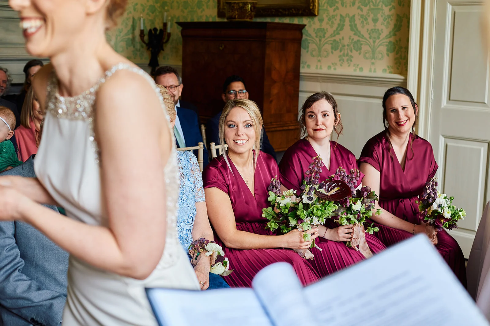 An over-the-shoulder view captures a joyful ceremony as bridesmaids in burgundy satin dresses sit holding bouquets while watching the proceedings.