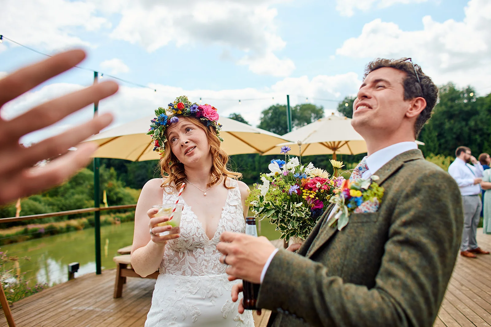 A joyful bride wearing a colourful floral crown and groom in a tweed jacket enjoy drinks and laughter outside during a summer wedding reception.