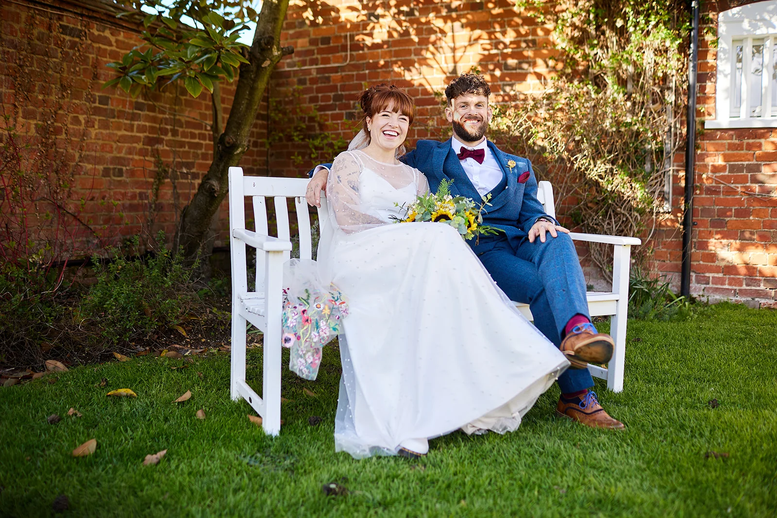 A smiling bride and groom sit together on a white wooden garden bench outdoors in front of a red brick wall.