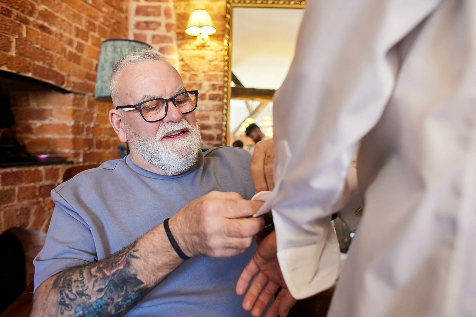 A mature groom with tattoos and glasses adjusts his cufflinks in a room with exposed brick walls during morning preparations.