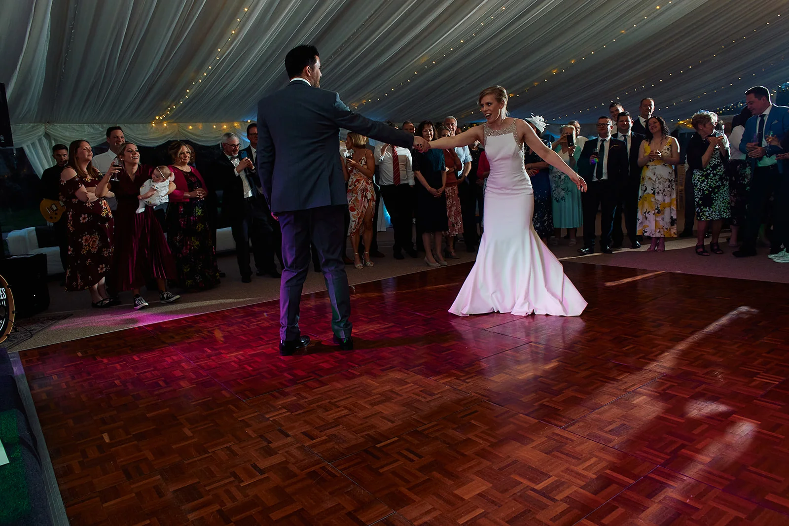 The newly married couple share their first dance on a polished wooden dance floor inside a marquee draped in fabric and fairy lights, surrounded by cheering wedding guests.