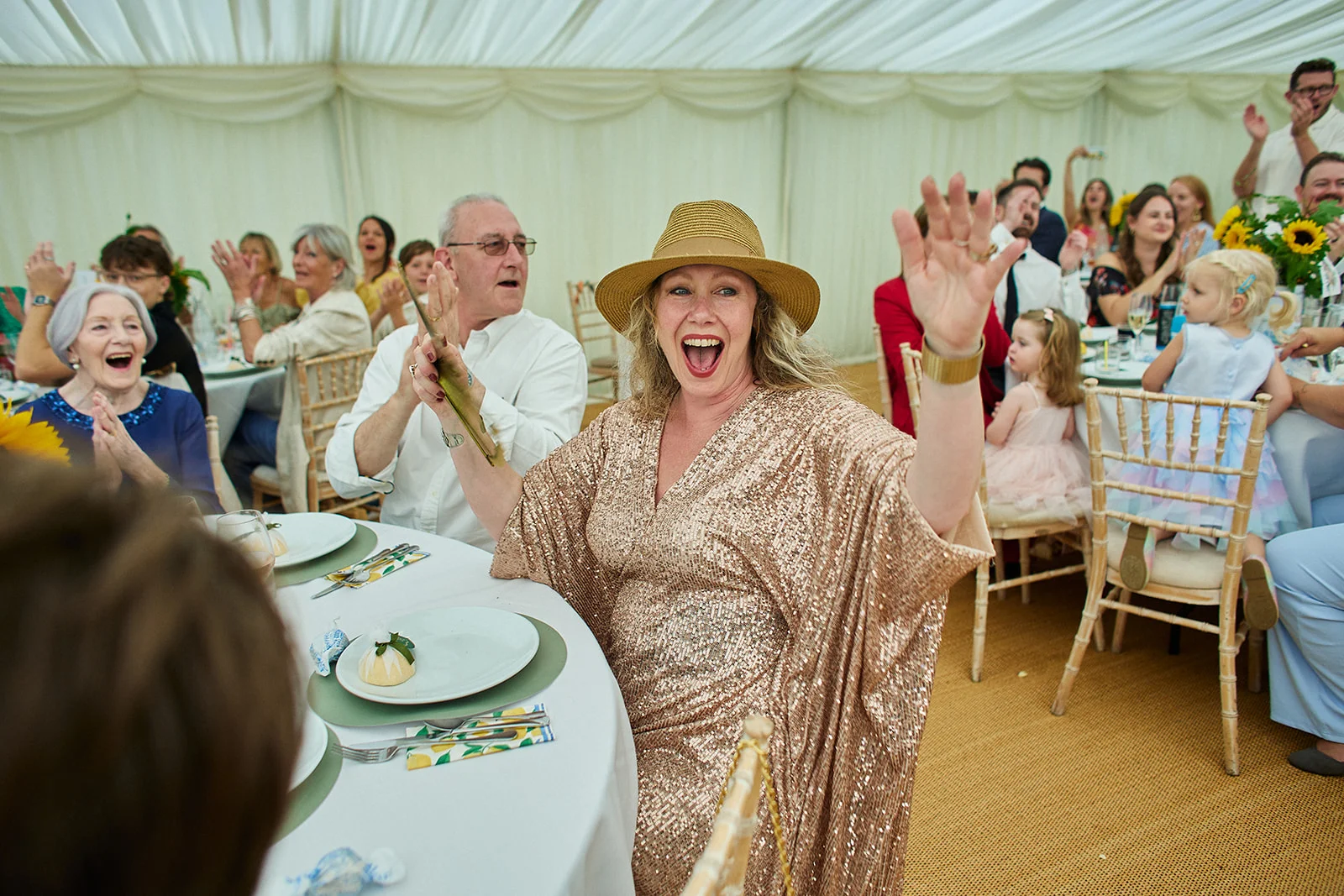 A joyful guest in a sparkling gold sequin dress and straw hat waves her hand excitedly while seated at a reception table inside a bright marquee.