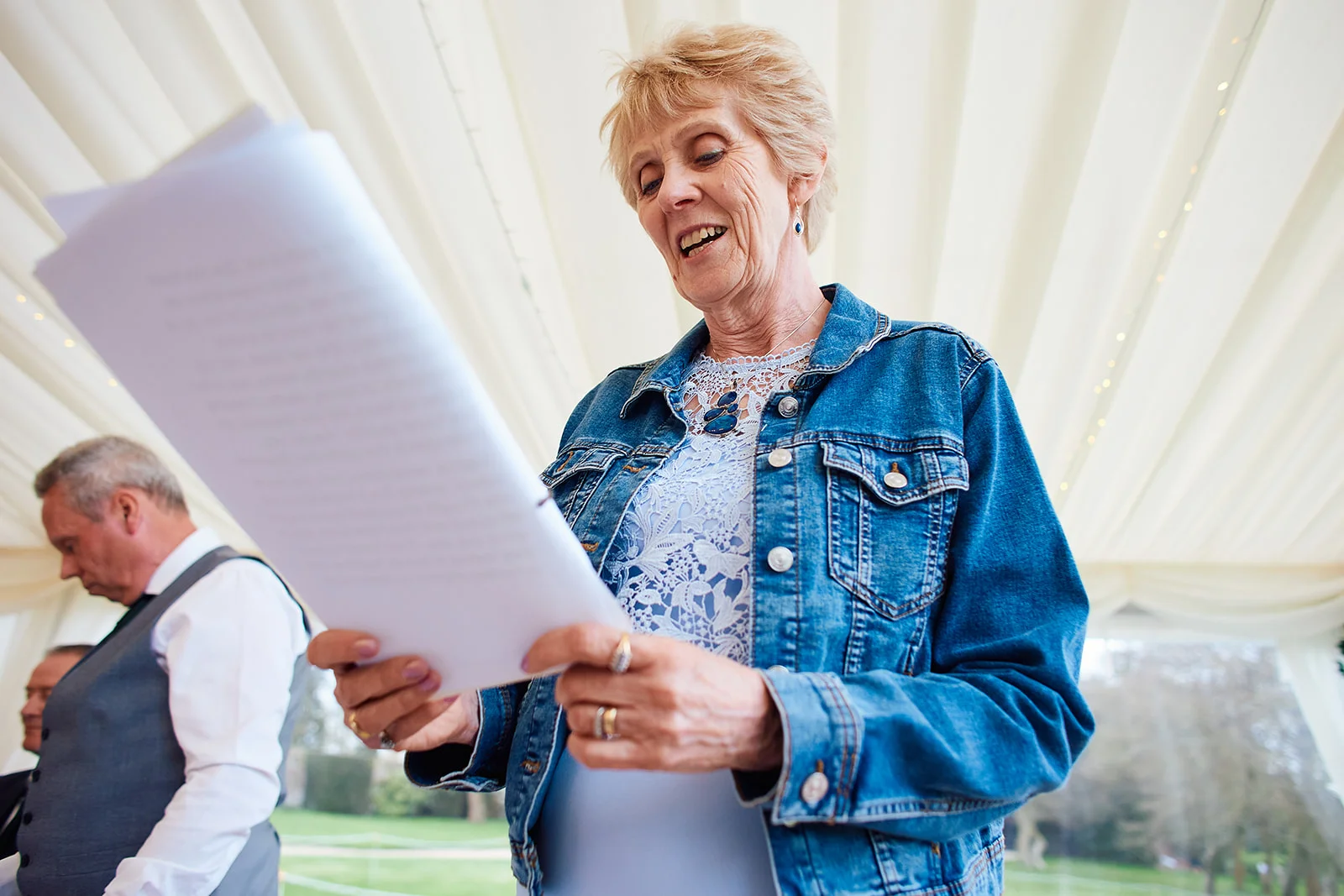 A mature guest happily reads from a speech document while wearing a denim jacket inside a marquee wedding reception.