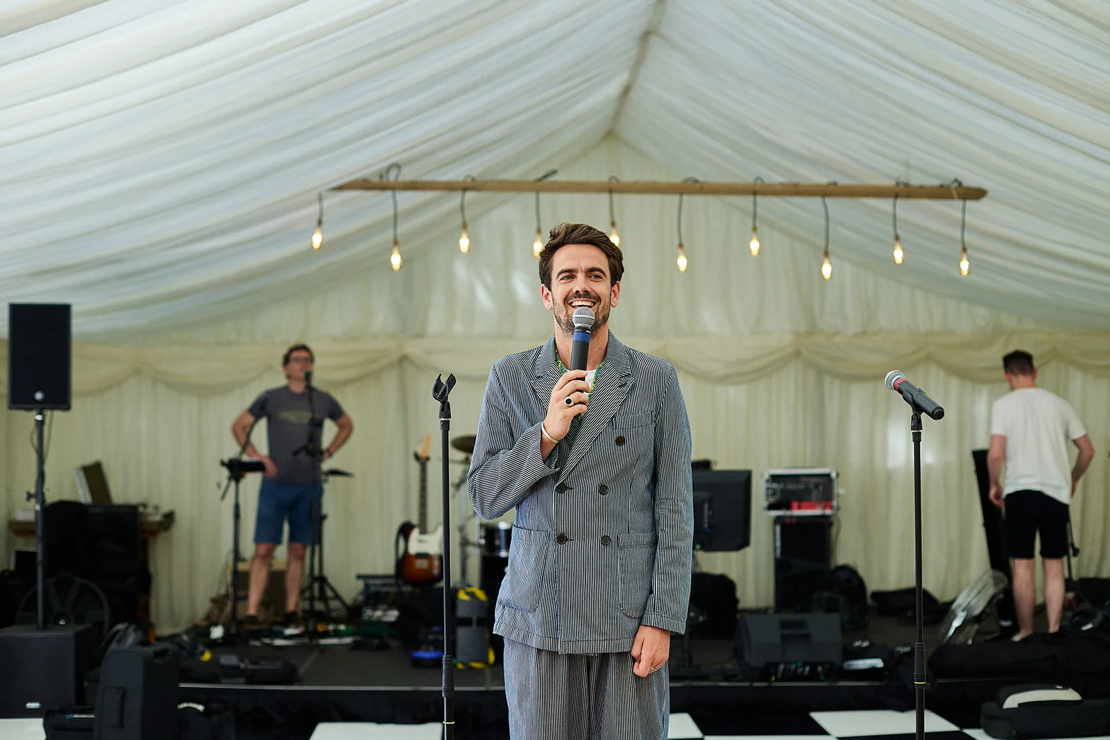 A smiling man in a striped double breasted suit delivers a speech into a microphone inside a draped marquee with stage lighting.