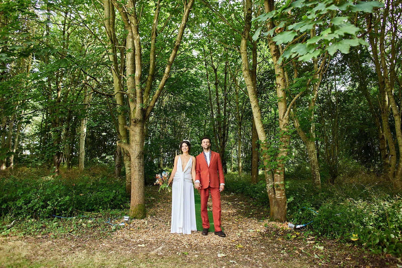 A newlywed couple holds hands and stands together along a green path winding through a lush, tree-filled woodland.
