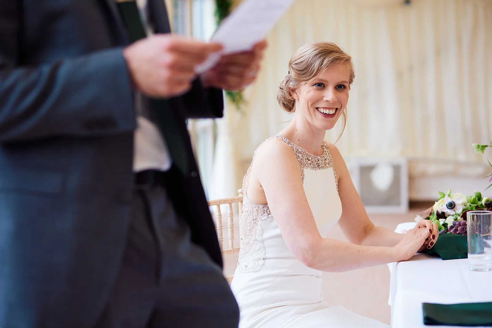 A smiling bride seated at the top table listens attentively while someone in the foreground holds speech notes.