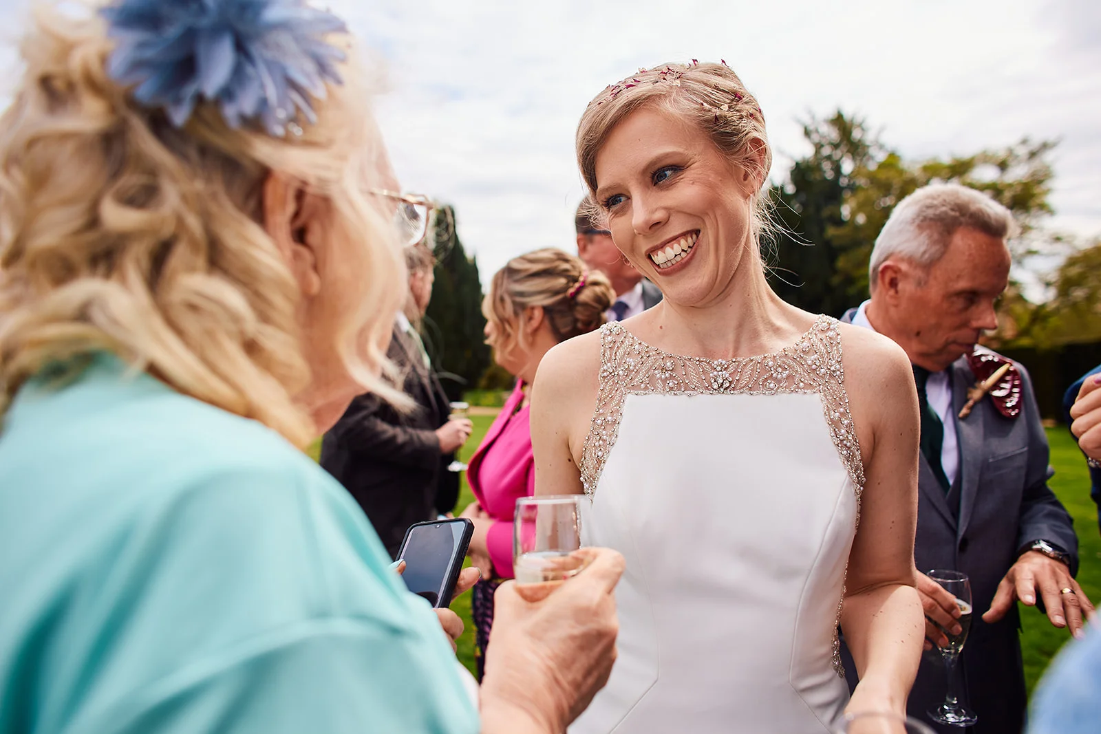 A smiling bride wearing an embellished high-neck gown chats with guests holding champagne glasses during an outdoor drinks reception.