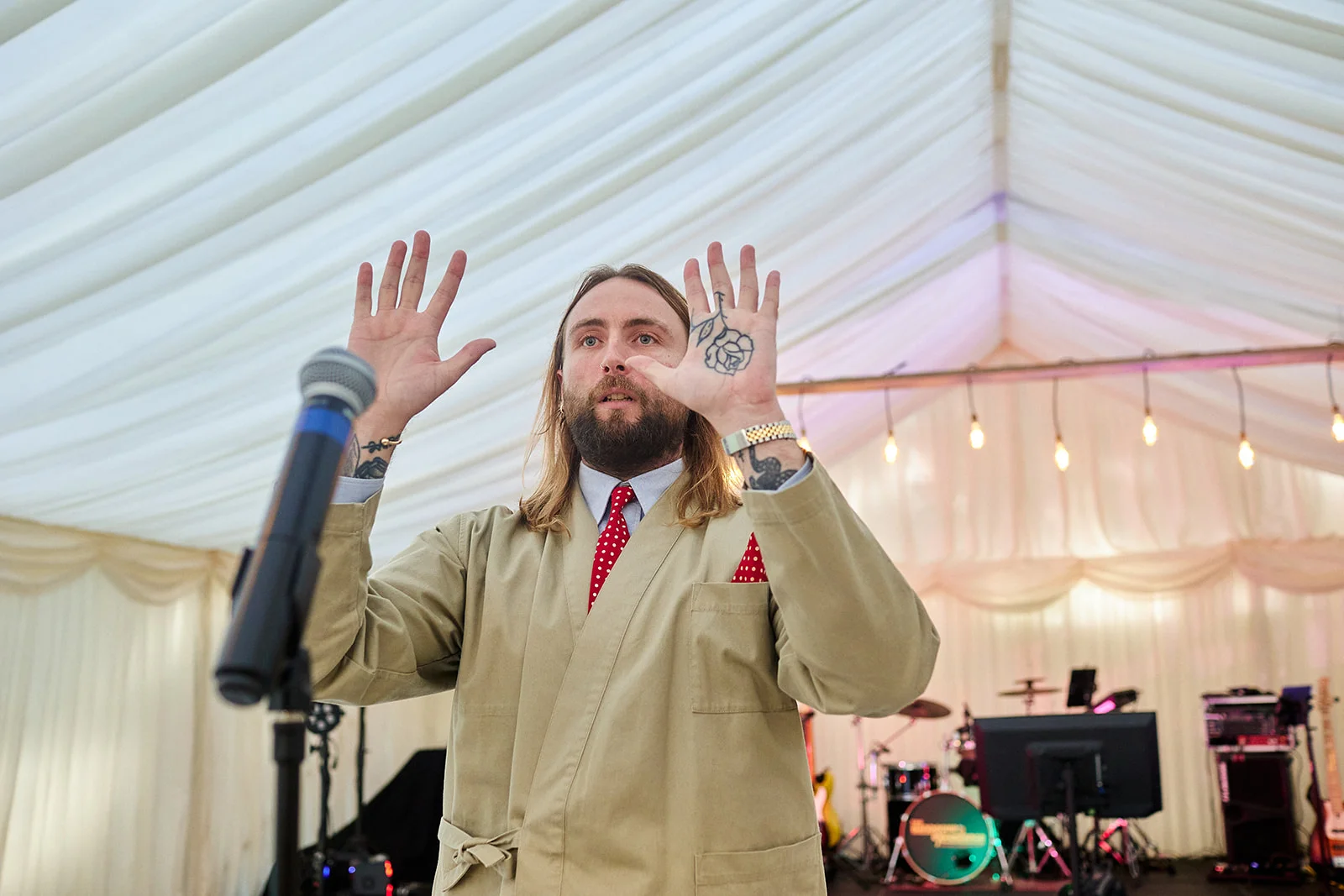 A bearded groom with long hair and palm tattoos gestures with both hands while giving a speech in a draped marquee during the wedding reception.