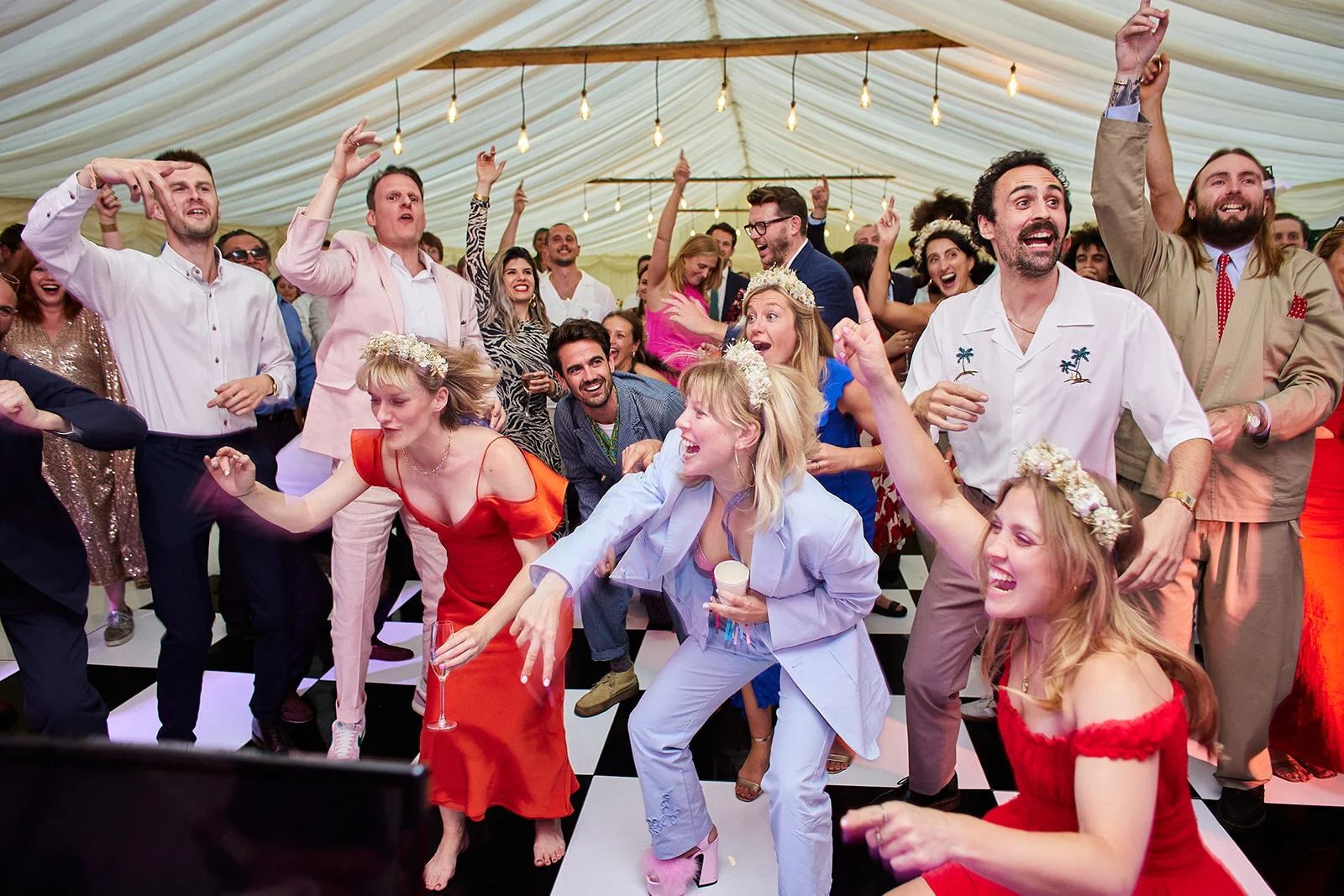 A joyous crowd of wedding guests dances enthusiastically on a black and white checkerboard dance floor beneath a draped marquee lit by festoon lights.