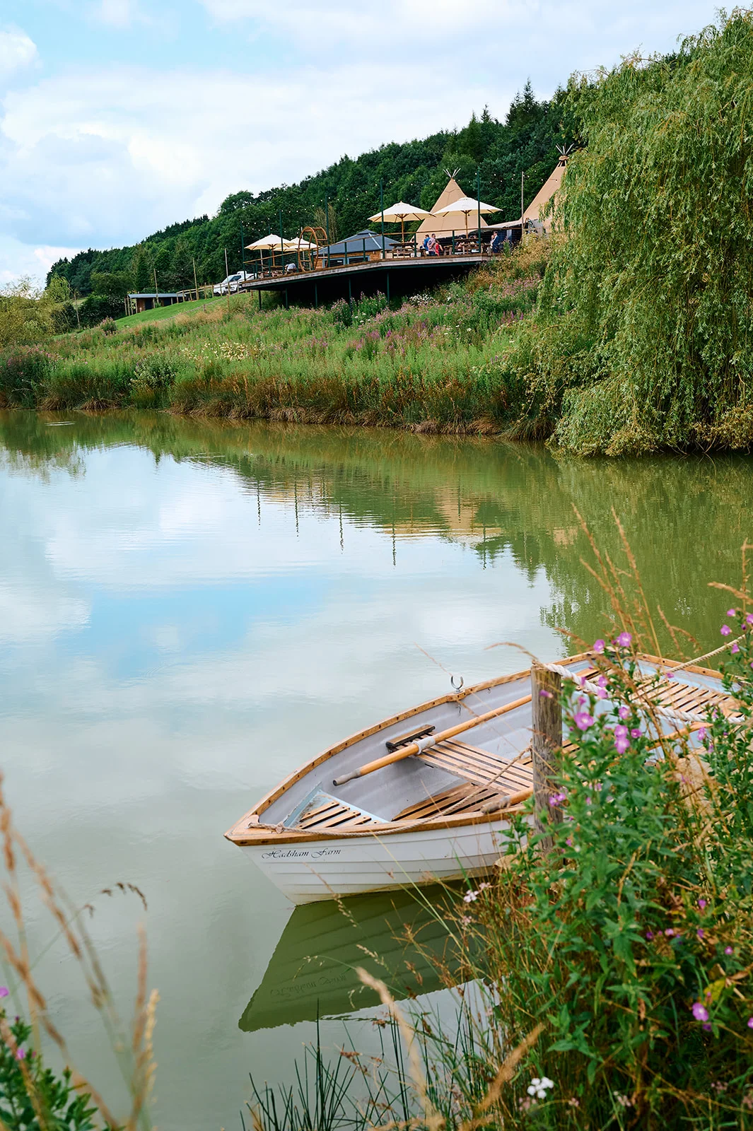 A rustic wooden rowing boat is moored by the edge of a tranquil lake, with a tipi wedding venue and green hillside visible in the background.
