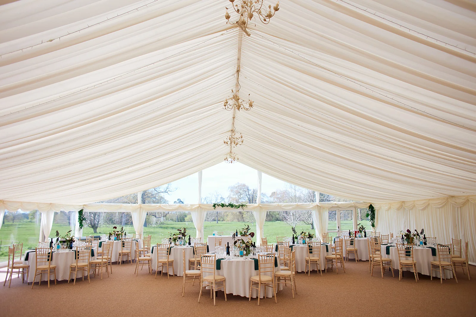 A marquee wedding reception venue features elegantly draped white ceilings, hanging chandeliers, and round tables set with white linens and green accents overlooking a countryside view.