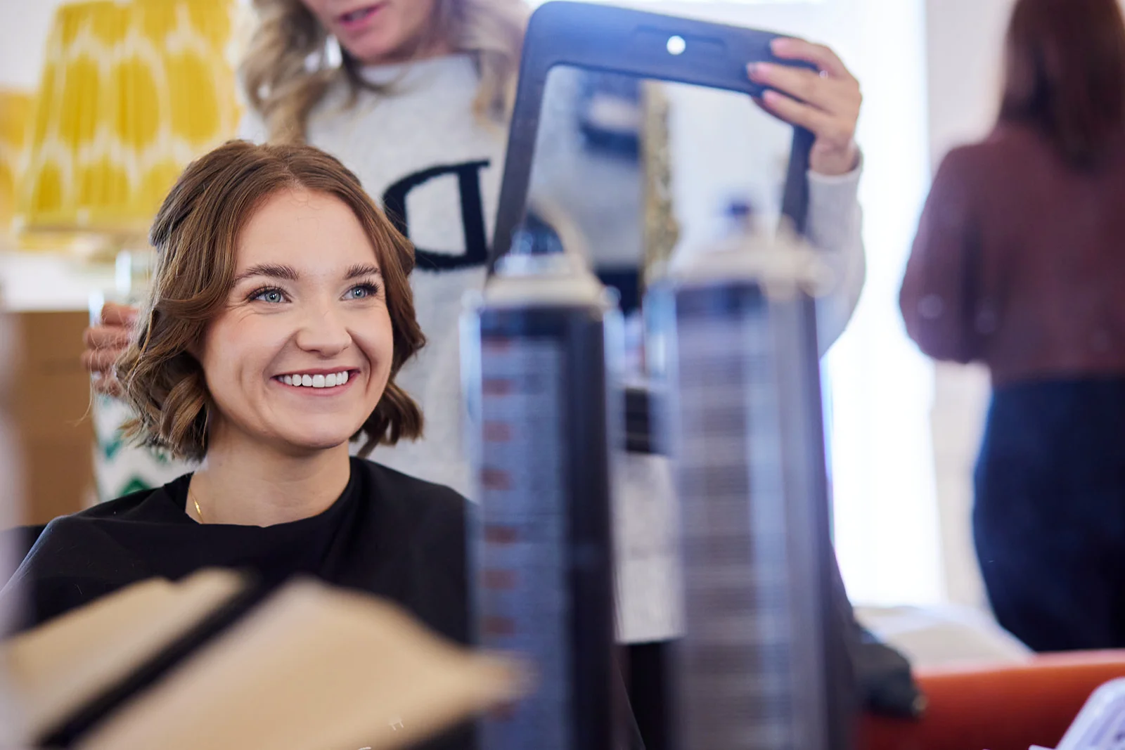 A smiling bride looks into a mirror while getting her hair styled during morning wedding preparations.