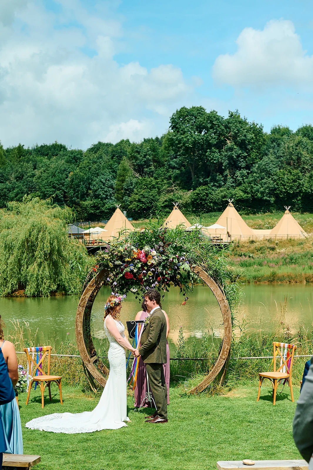 A bride and groom exchange vows during an outdoor lakesidic ceremony beneath a large circular floral arch, with tipis visible in the background.