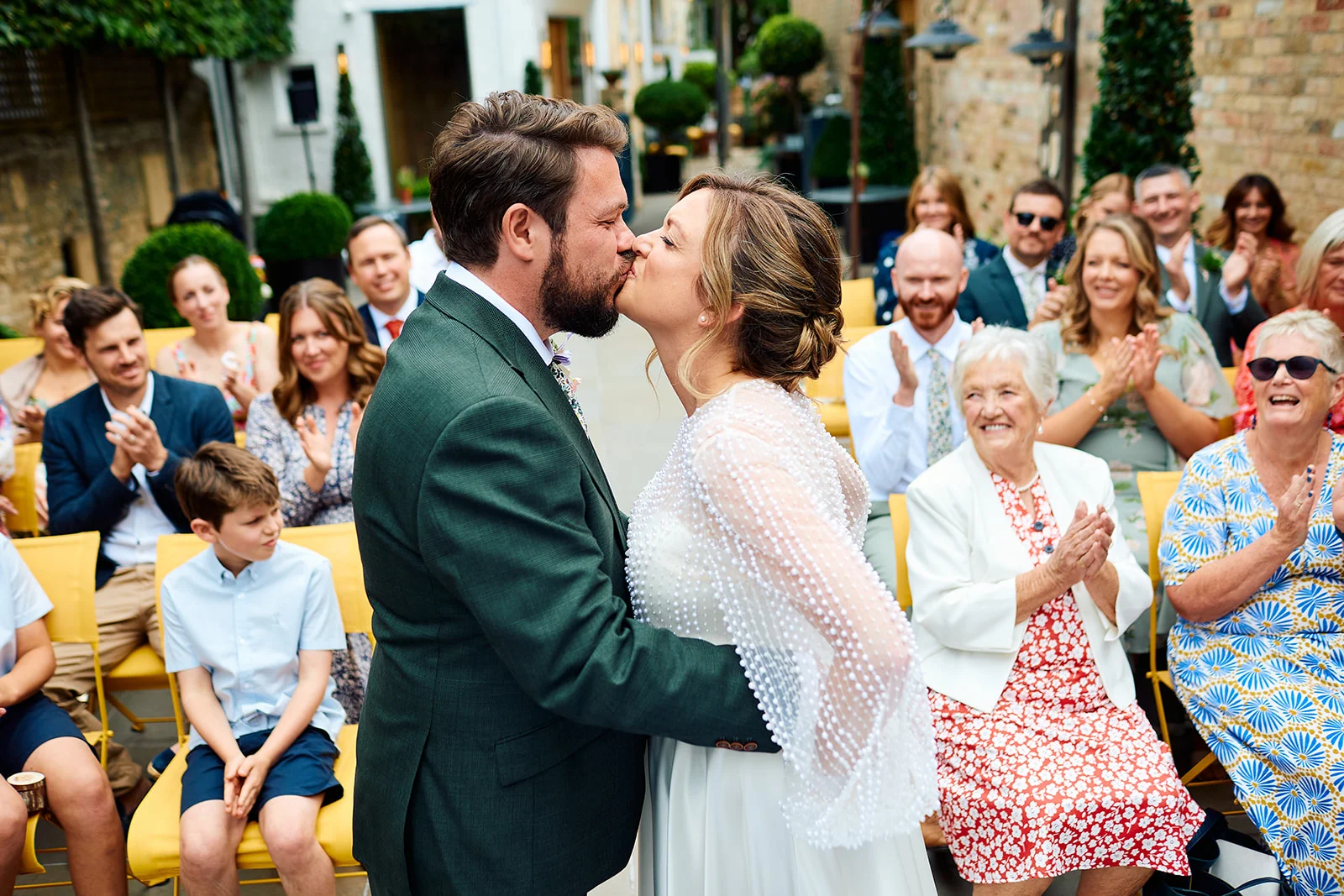 Newlyweds share their first kiss during an outdoor courtyard ceremony while family and friends applaud and celebrate.