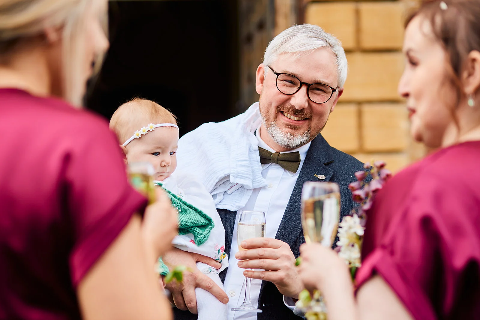 A smiling guest wearing glasses and a bow tie holds a baby while mingling and holding a glass of champagne during the wedding drinks reception.
