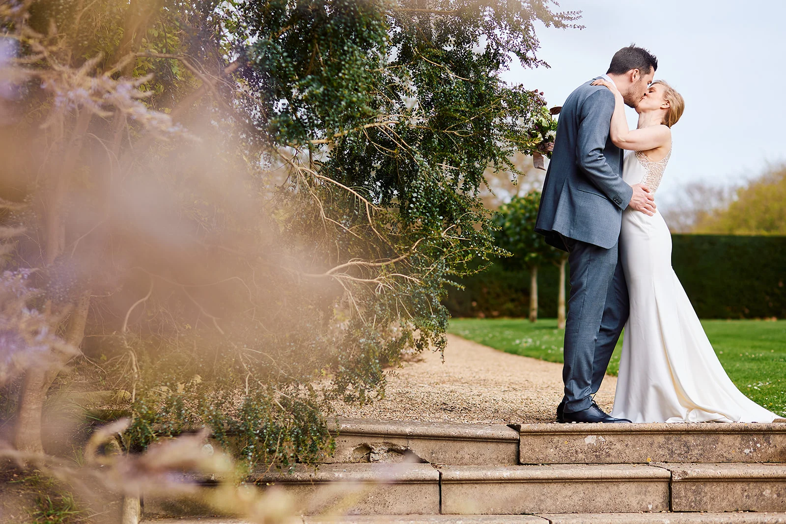 A newlywed couple shares a tender kiss while standing on stone steps framed by lush garden foliage and trees.