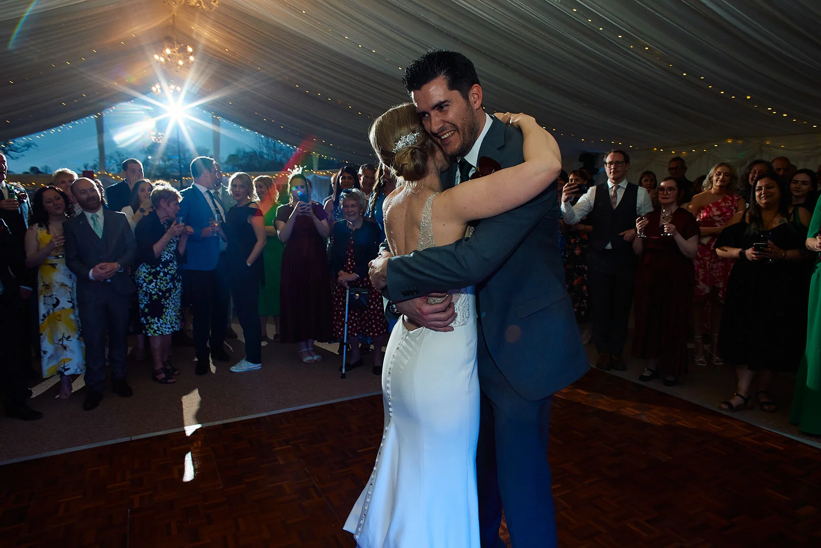 A newlywed couple share an emotional embrace during their first dance on a wooden dance floor inside a lit marquee with cheering guests watching.