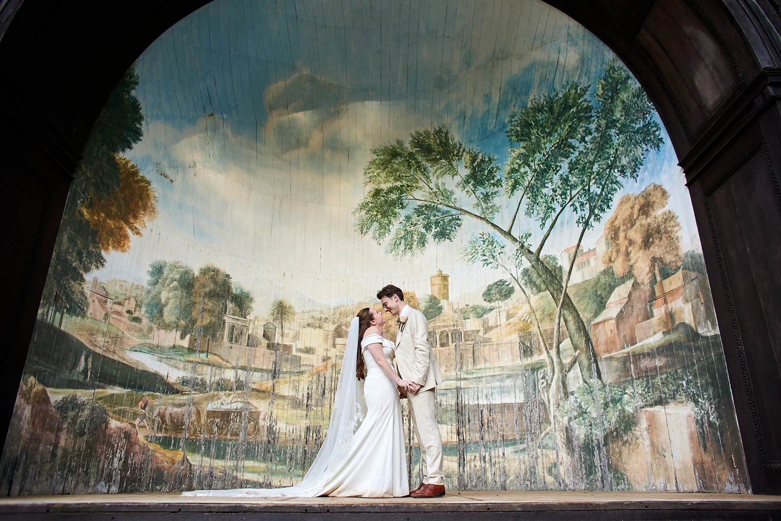 A bride and groom stand hand in hand inside a grand arched alcove featuring a large painted mural of a landscape behind them.