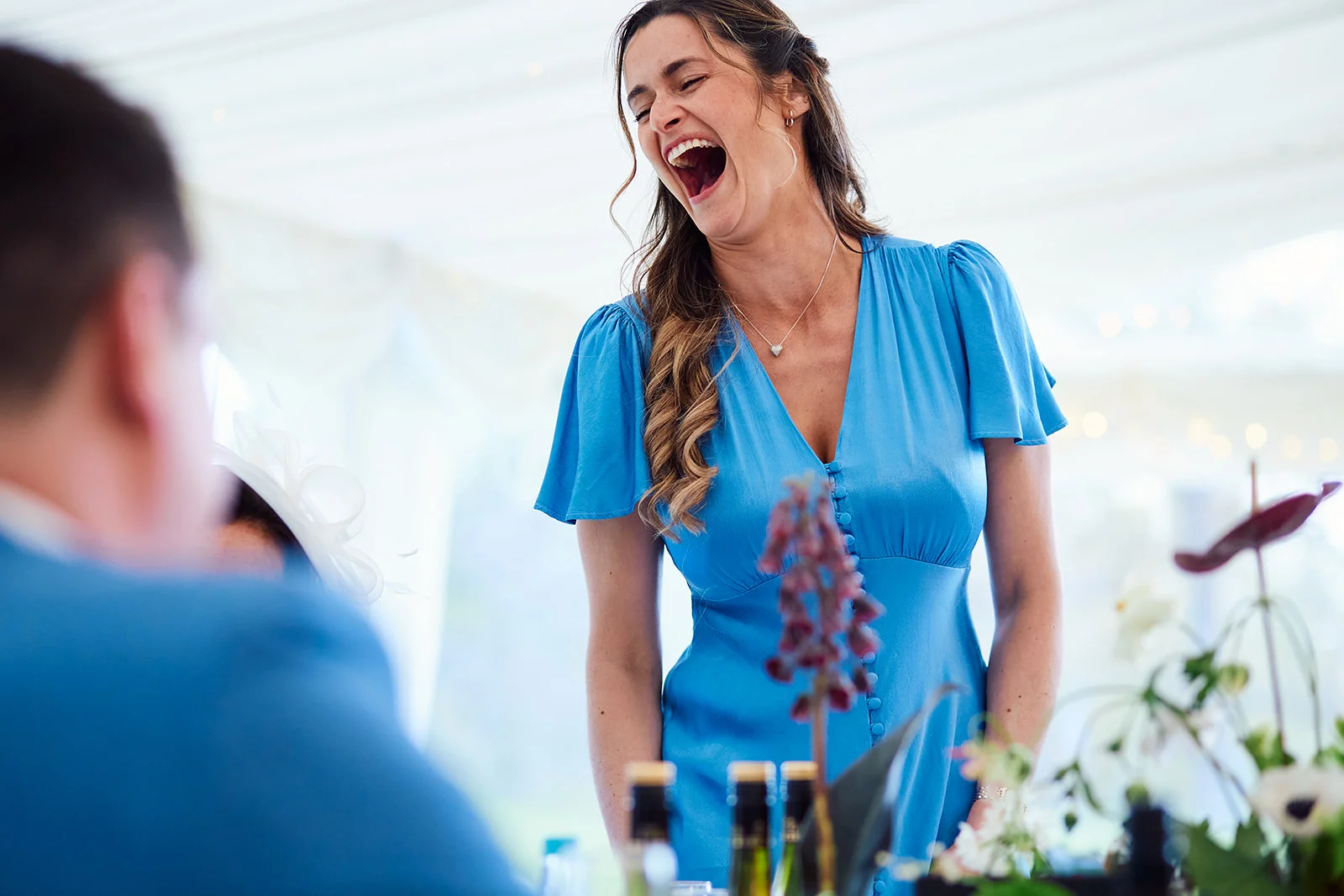 A guest in a blue dress laughs joyfully during the wedding speeches inside a bright marquee reception.