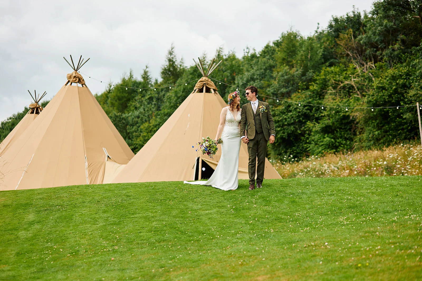 A newlywed couple walks hand in hand across a green lawn outside large canvas tipis framed by trees.