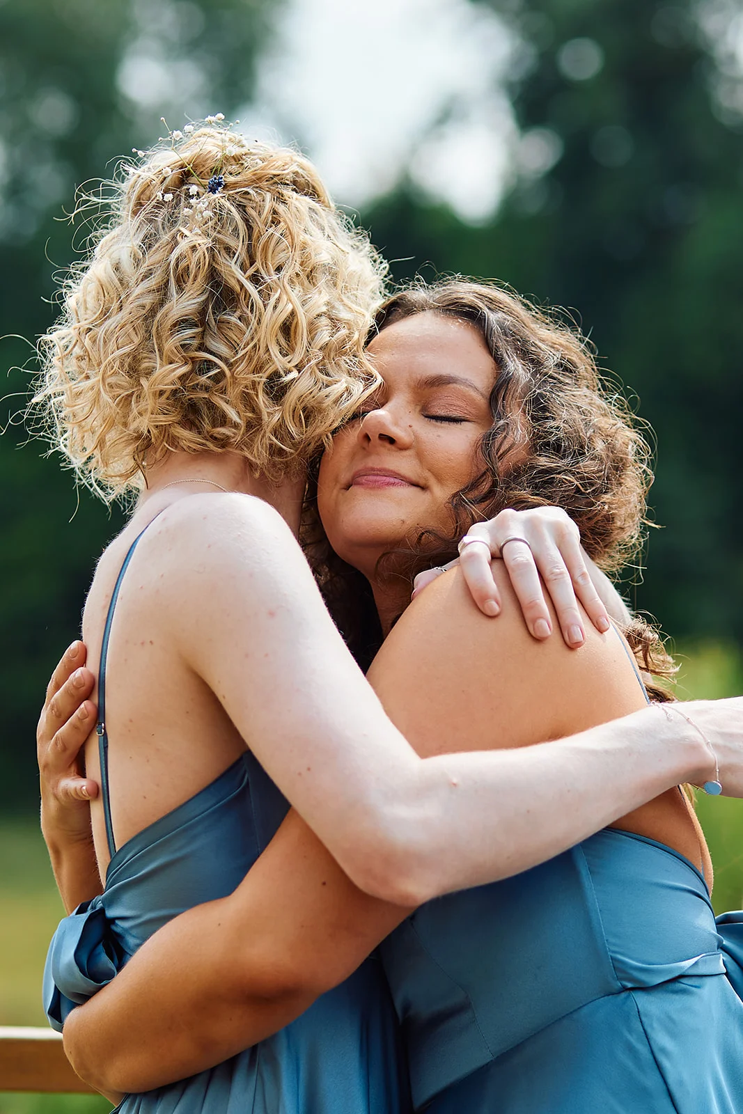 Two brides with curly hair in blue dresses share a tender, emotional embrace outdoors.