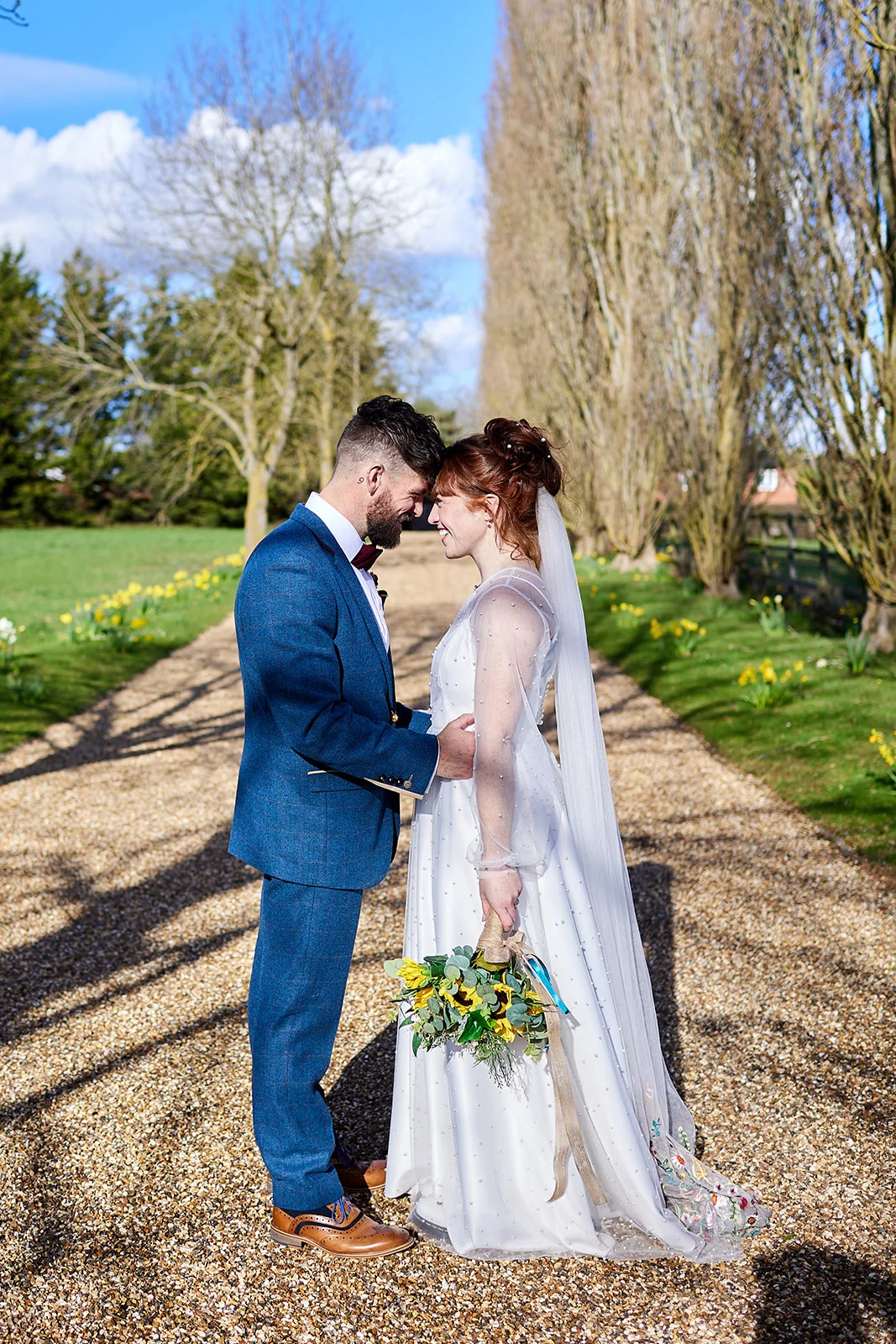 A newlywed couple shares a tender forehead kiss on a sunlit gravel path lined with trees and yellow spring flowers.