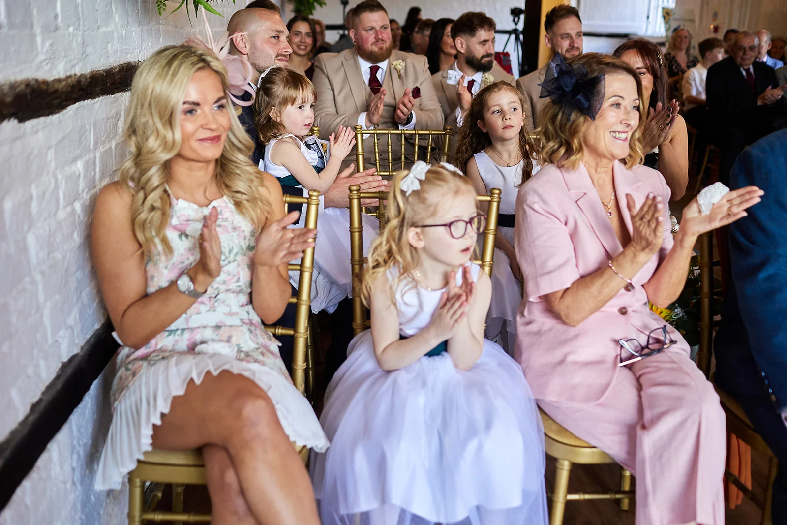 Wedding guests happily applauds while seated during the ceremony, including a woman in a pink suit and young flower girls in white dresses.