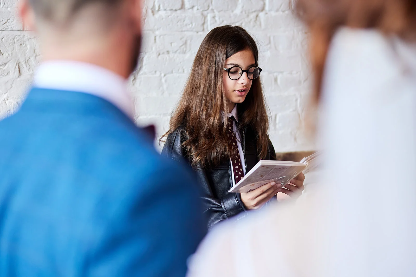 A young wedding guest wearing glasses and a black jacket reads through a booklet while framed by guests in the foreground.