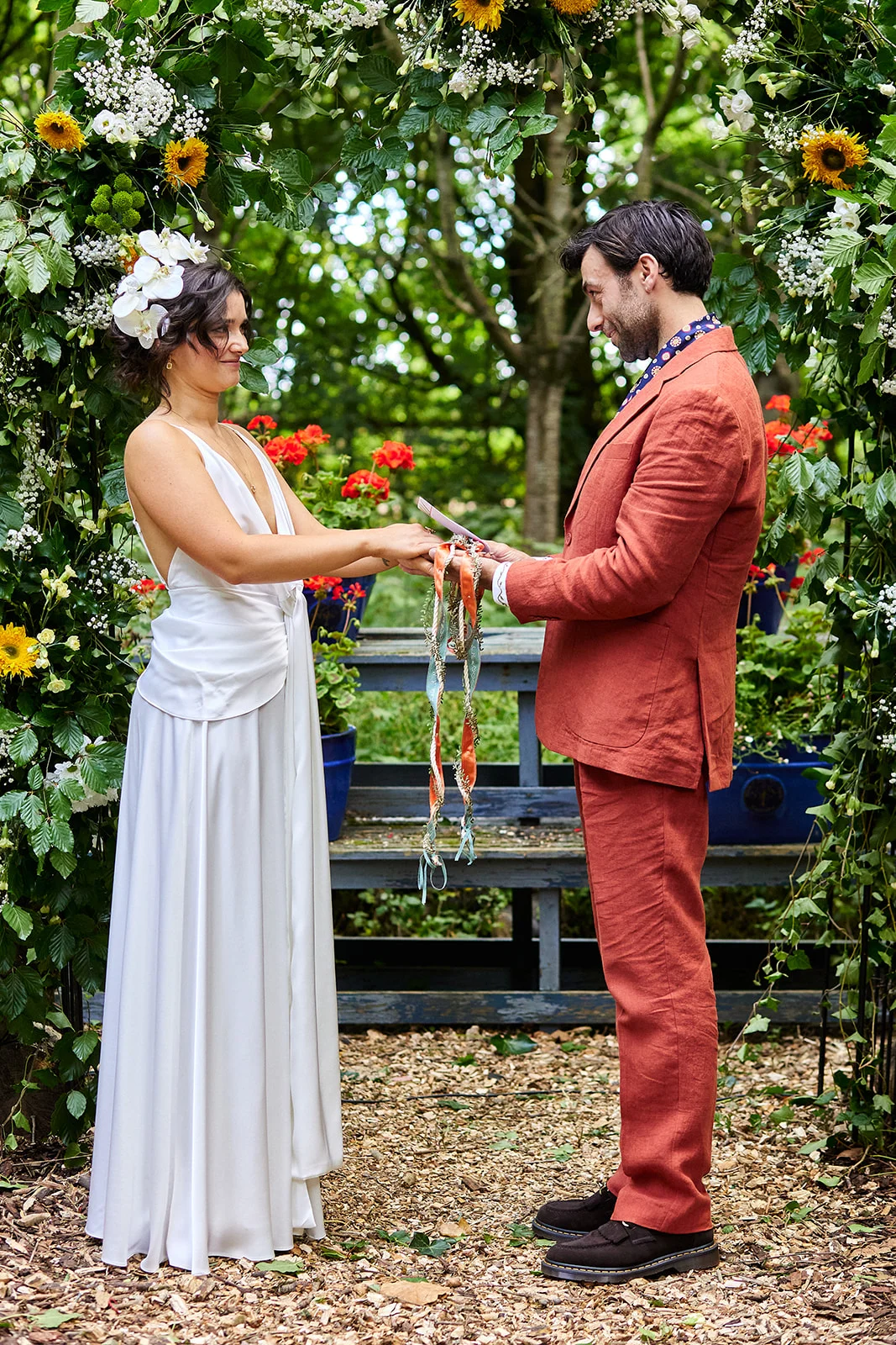 A bride and groom participate in an outdoor handfasting ceremony beneath a lush floral arch adorned with greenery and sunflowers in a woodland setting.
