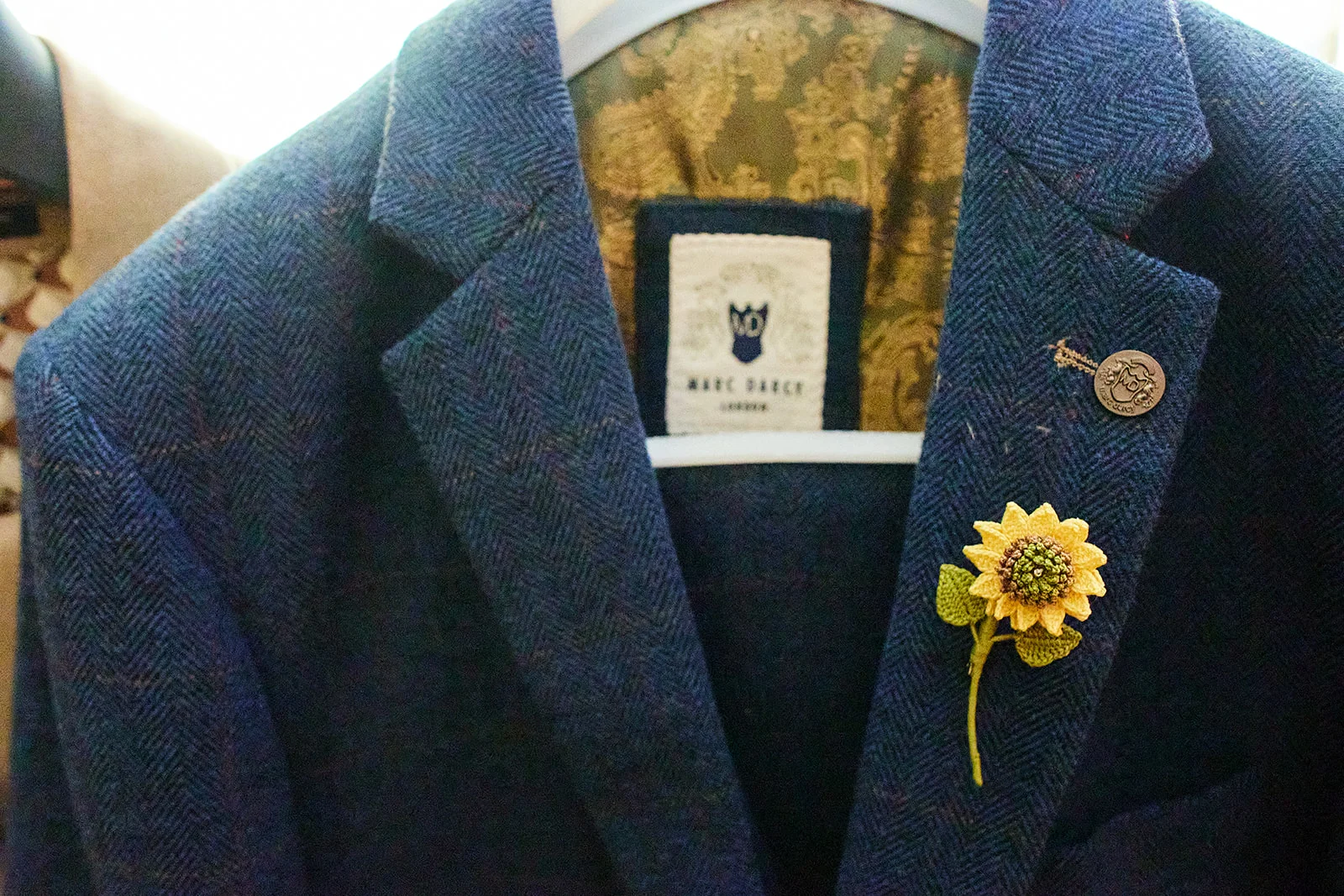 A tailored navy herringbone tweed suit hangs on a white hanger, featuring a decorative handmade sunflower lapel pin and a branded label inside.