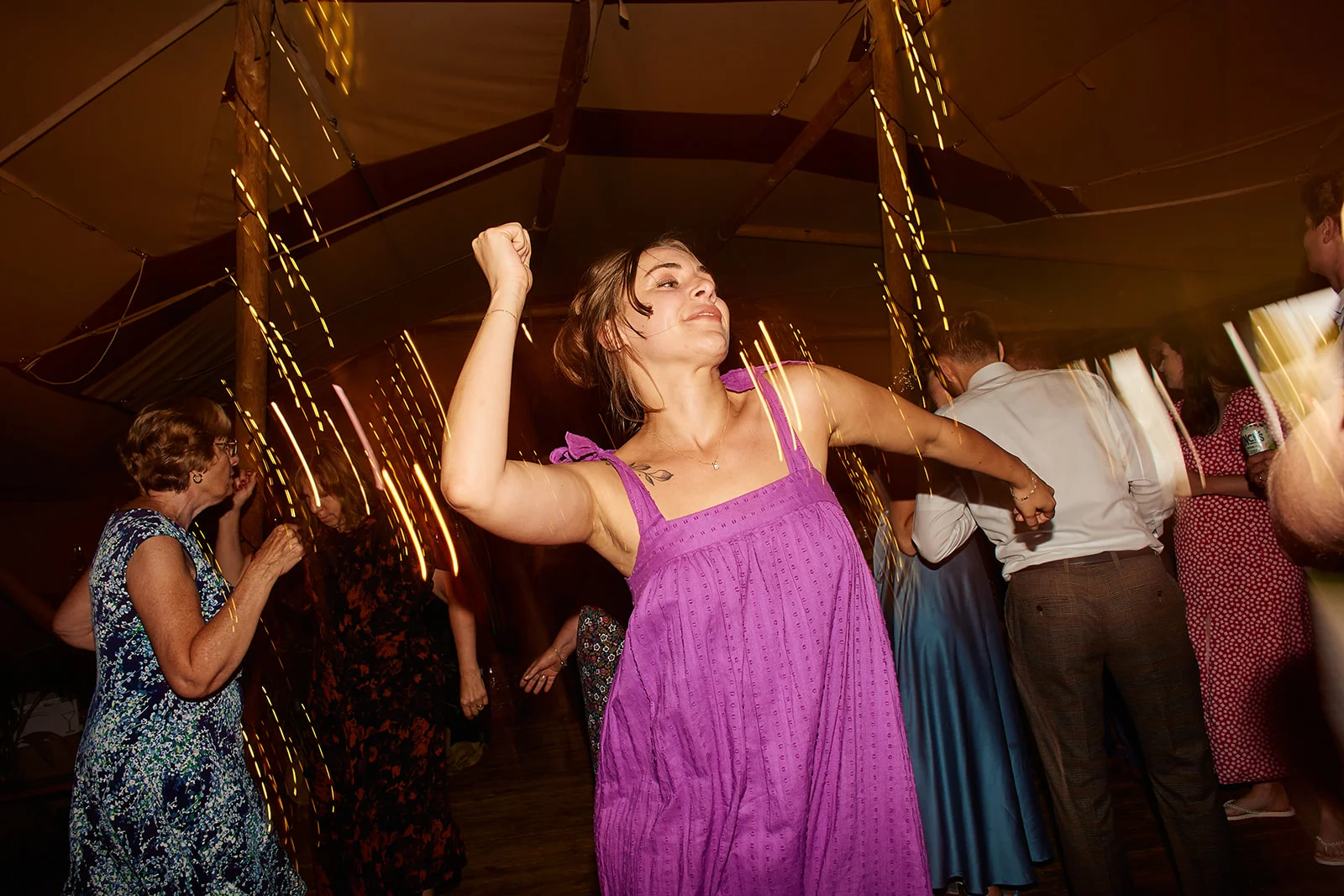 A guest in a vibrant purple dress dances energetically on a lively marquee dance floor under warm string lighting.