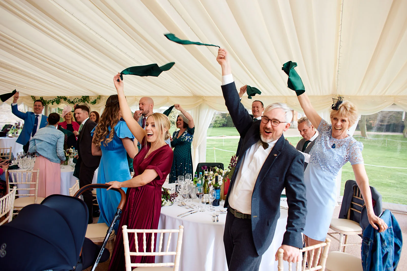 Wedding guests cheering and joyfully waving napkins in the air during the speeches inside a brightly lit marquee.