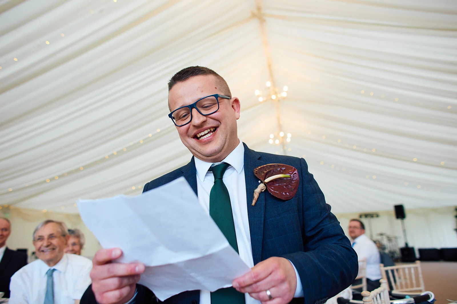 A smiling groom in a navy suit and green tie laughs heartily while reading his speech inside a draped marquee.