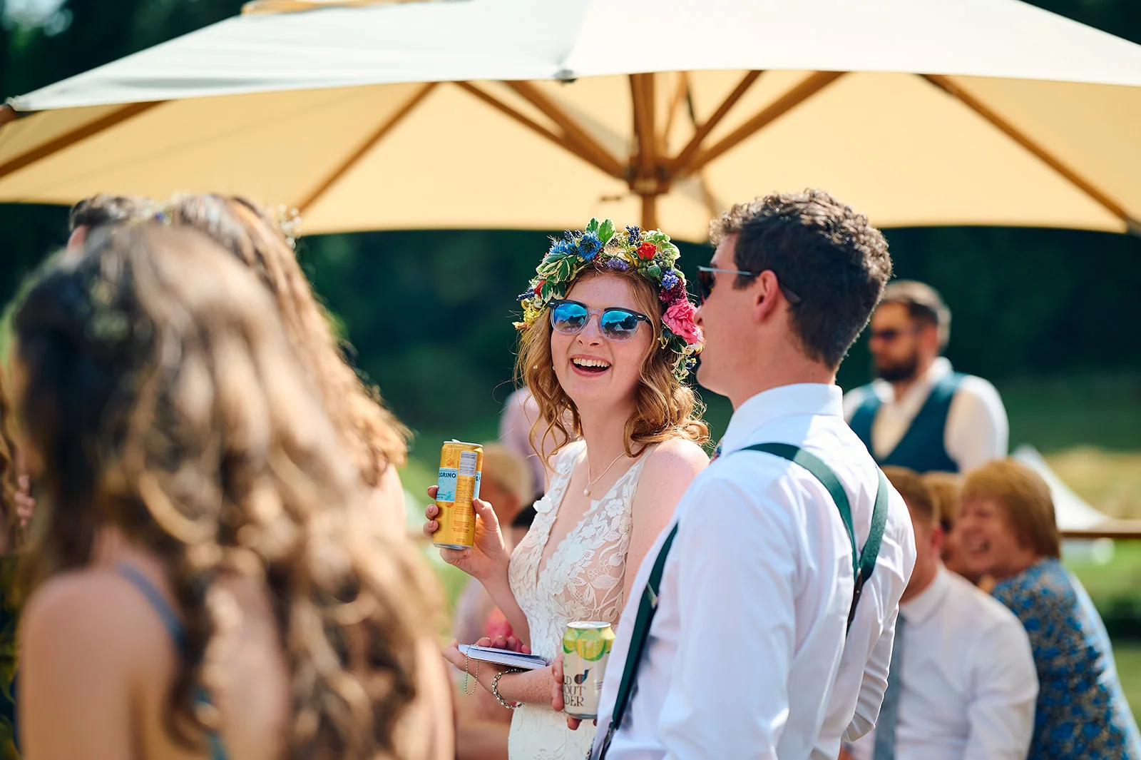 A laughing bride wearing a floral crown and sunglasses holds a drink next to a guest in a white shirt and braces during an outdoor drinks reception.