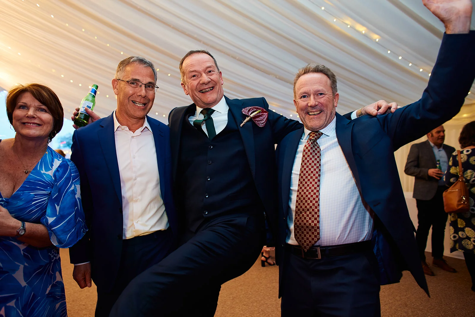 A joyful group of wedding guests share a lighthearted, laughing embrace under the draped ceiling of a marquee reception.