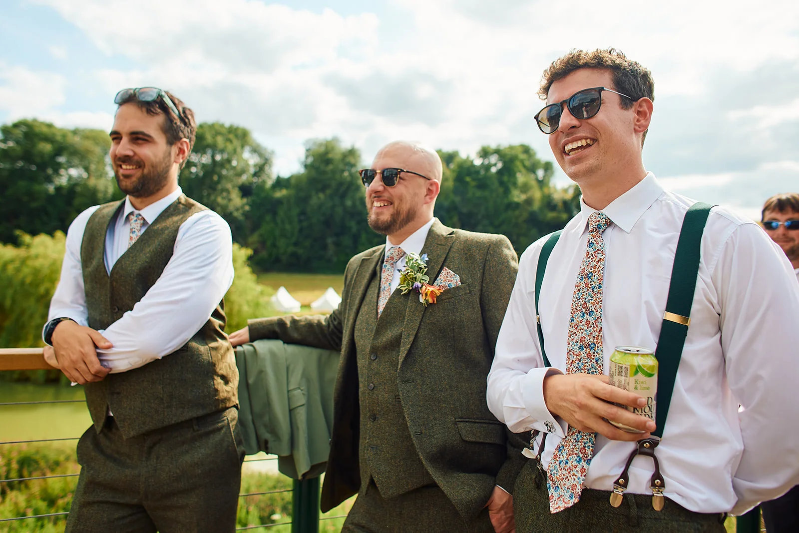 Three smiling groomsmen wearing sunglasses and tweed waistcoats stand outdoors during a sunny wedding reception, with one holding a drink.