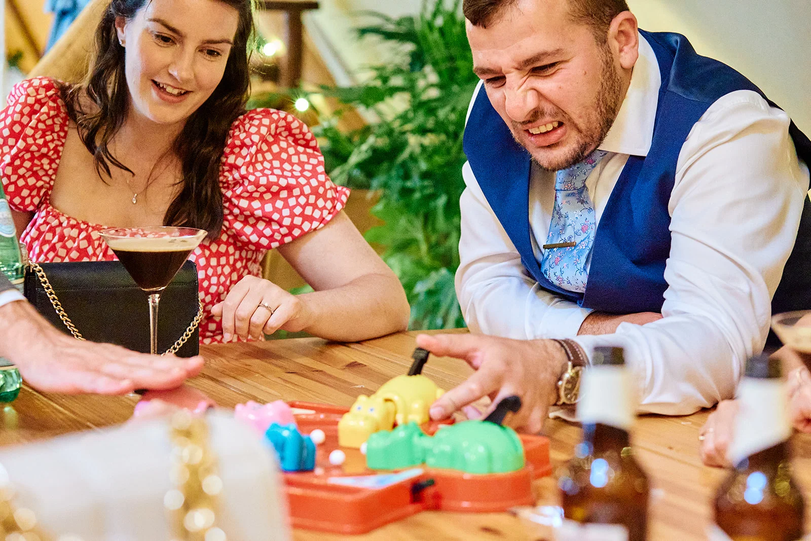 A guest laughing intently while playing a table game during a wedding reception, with a woman smiling beside him and a cocktail on the table.