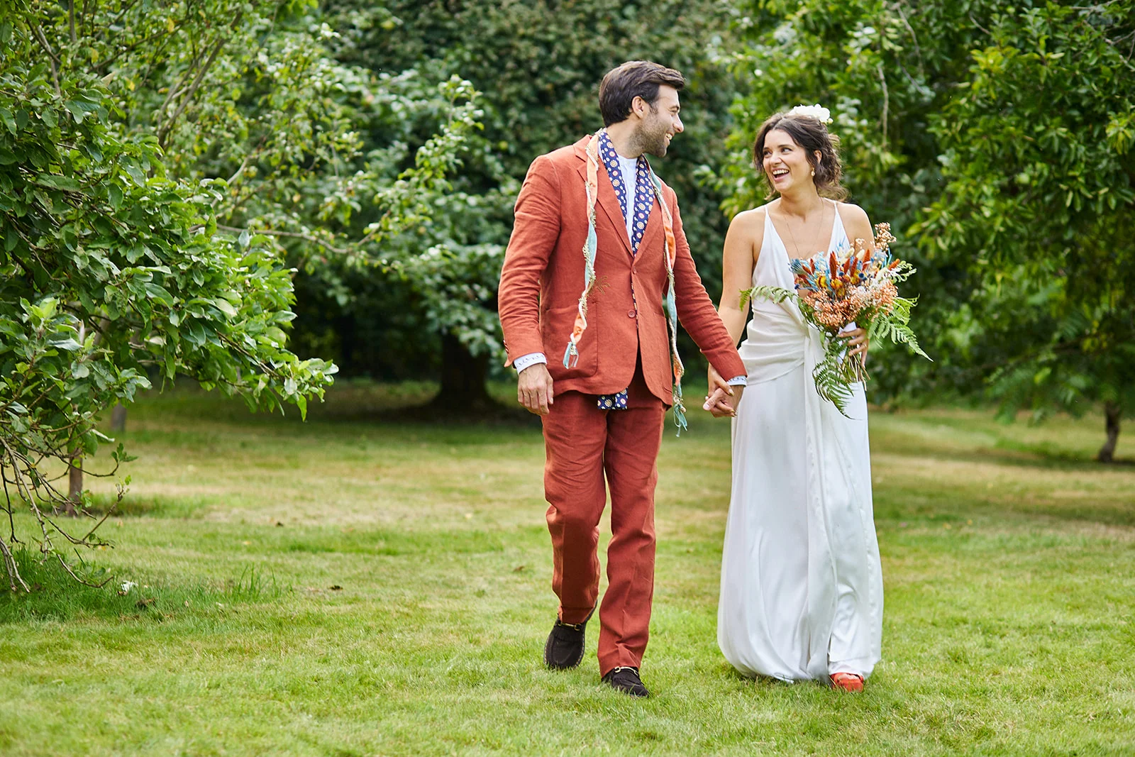 A joyful newlywed couple walks hand in hand through a lush orchard, with the bride holding a colourful bouquet and wearing a flowing slip dress while the groom sports a terracotta suit.