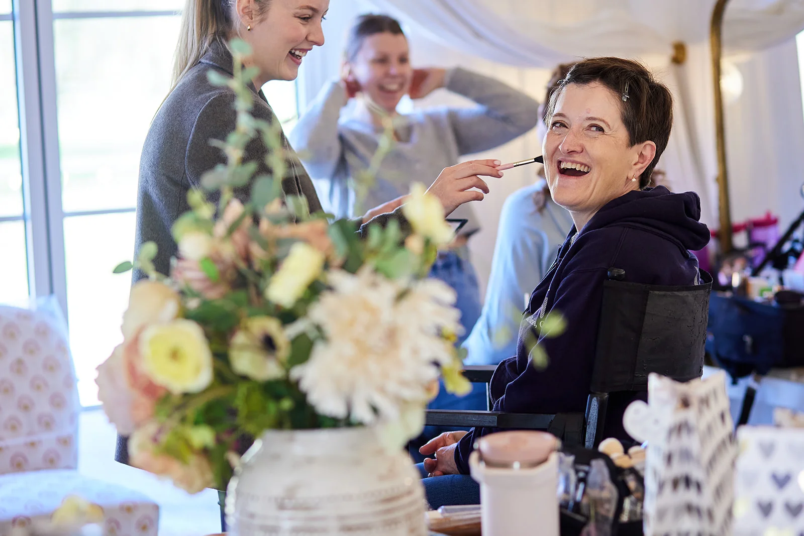 A bride laughs joyfully while having her makeup applied during the morning preparations surrounded by smiling friends and fresh floral arrangements.