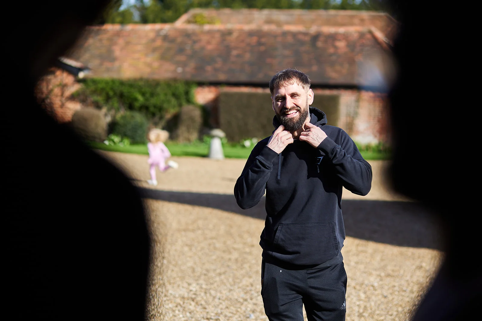 A smiling groom wearing a black hoodie stands outdoors in a gravel courtyard, framed by dark silhouettes in the foreground.