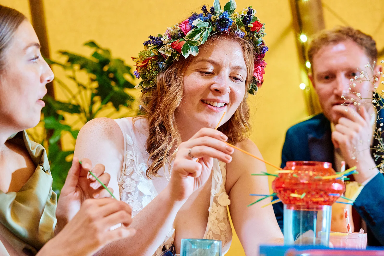 A smiling bride wearing a vibrant fresh flower crown plays a game at a wedding reception table surrounded by guests.