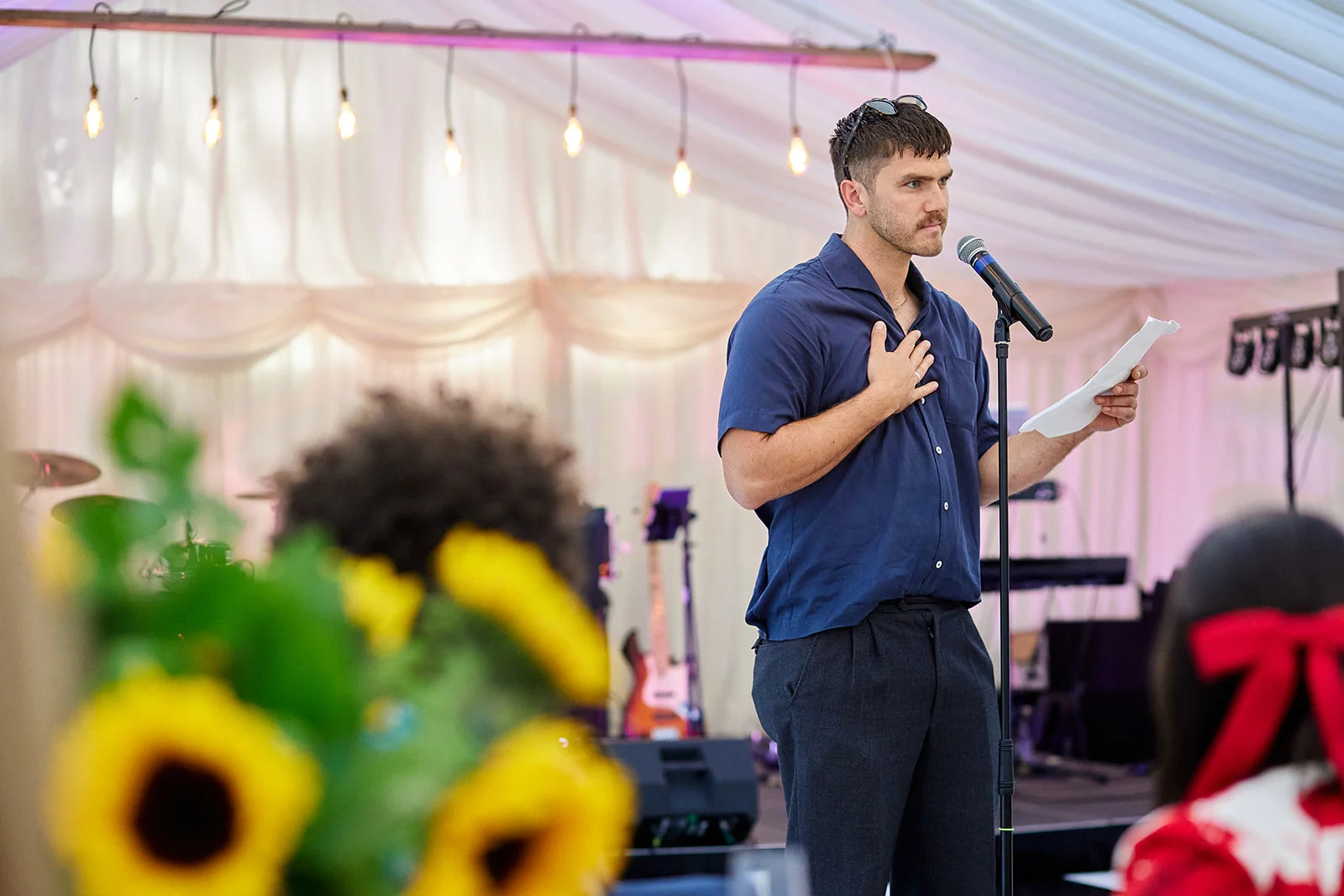 A groom delivers an emotional speech holding a microphone and notes inside a marquee decorated with hanging string lights and vibrant yellow sunflowers in the foreground.