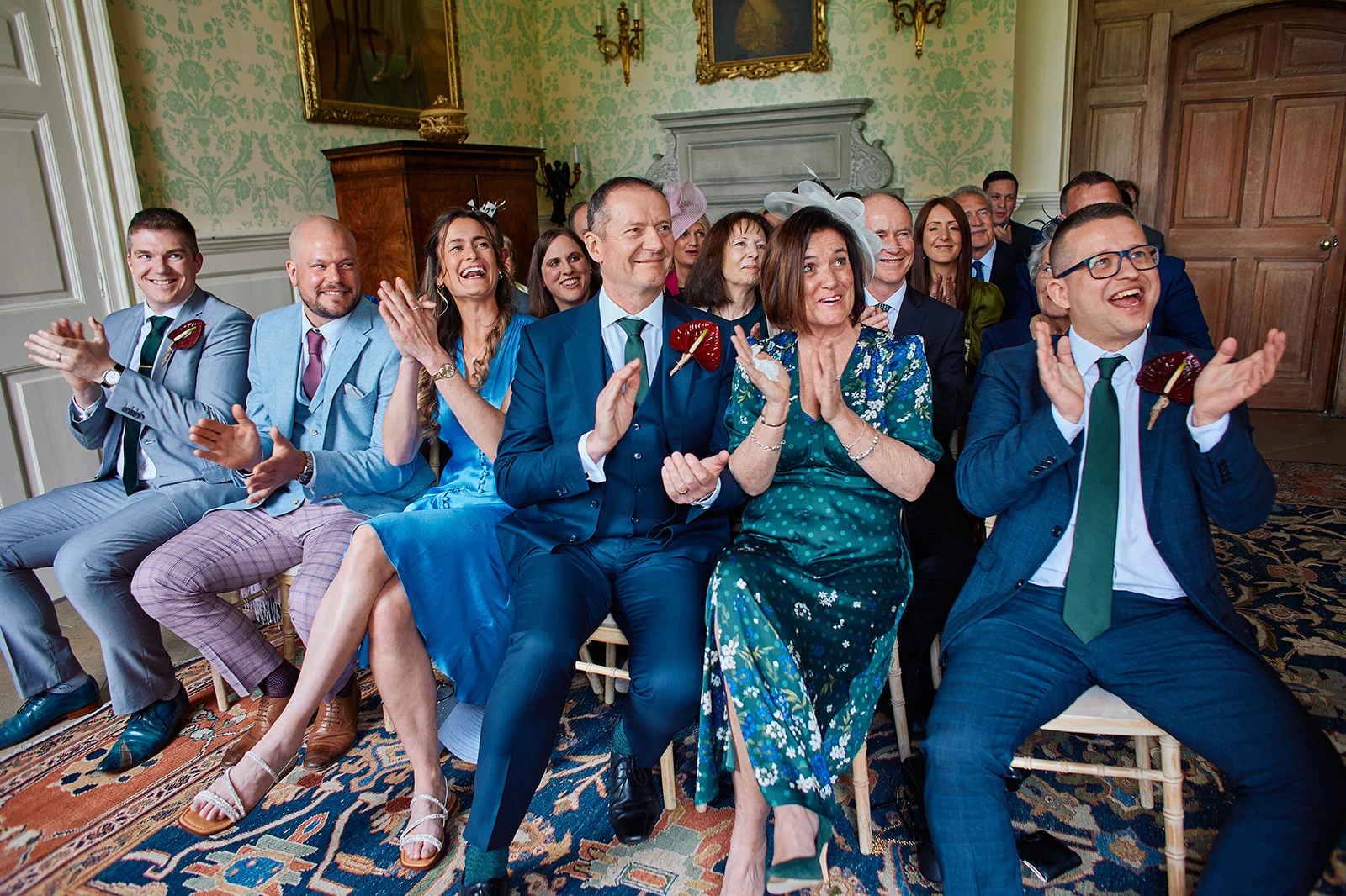 A joyful group of wedding guests seated in a grand manor house ballroom applaud enthusiastically during a ceremony.