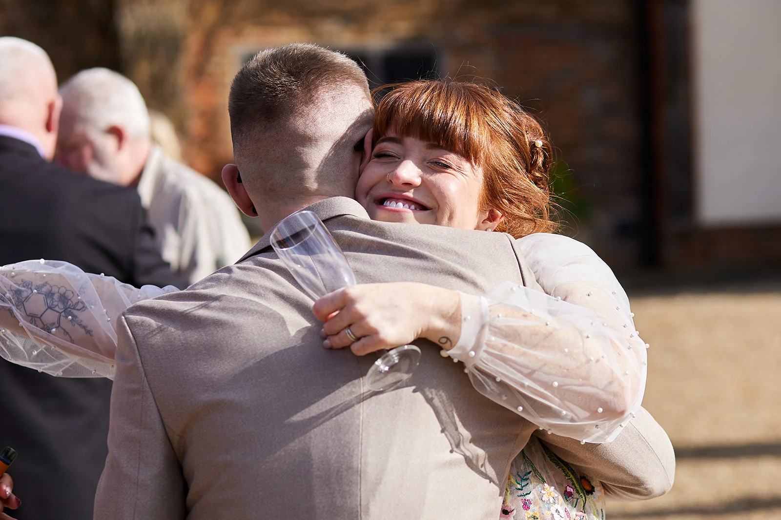A joyful red-haired bride with a fringe and pearl-detailed sheer sleeves warmly embraces her groom in a beige suit while holding a champagne flute outdoors.