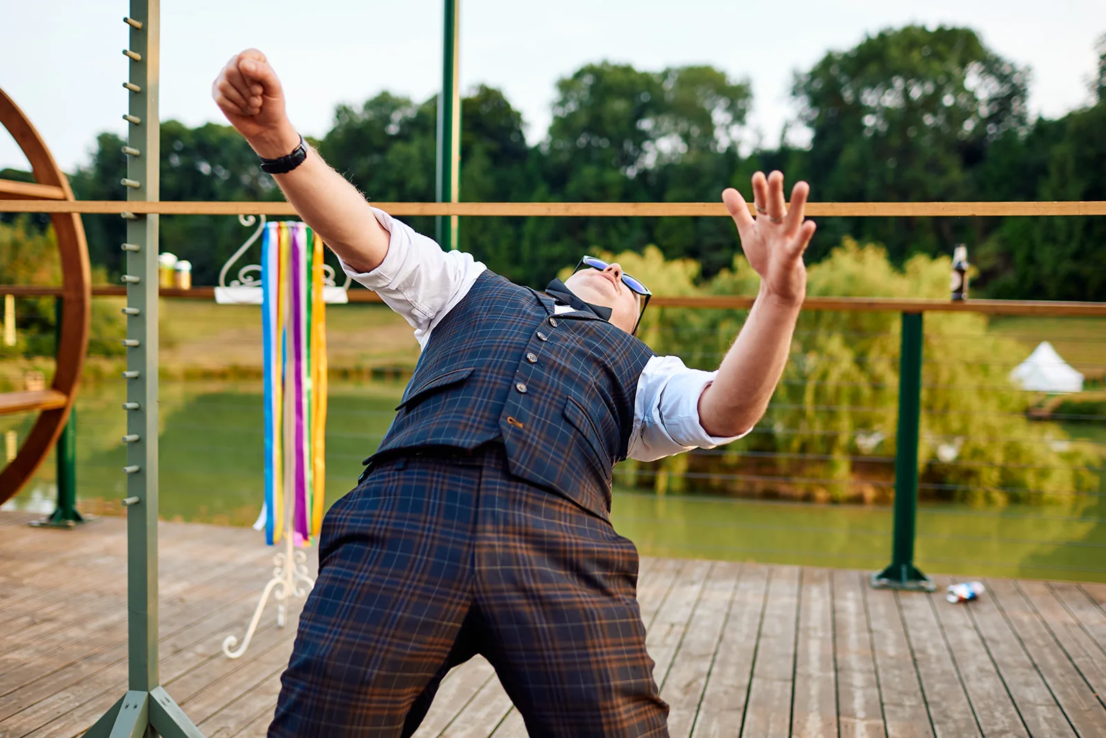 A groom in a checked waistcoat and sunglasses leans back dramatically with his arms raised in celebration on a wooden deck by a lake.