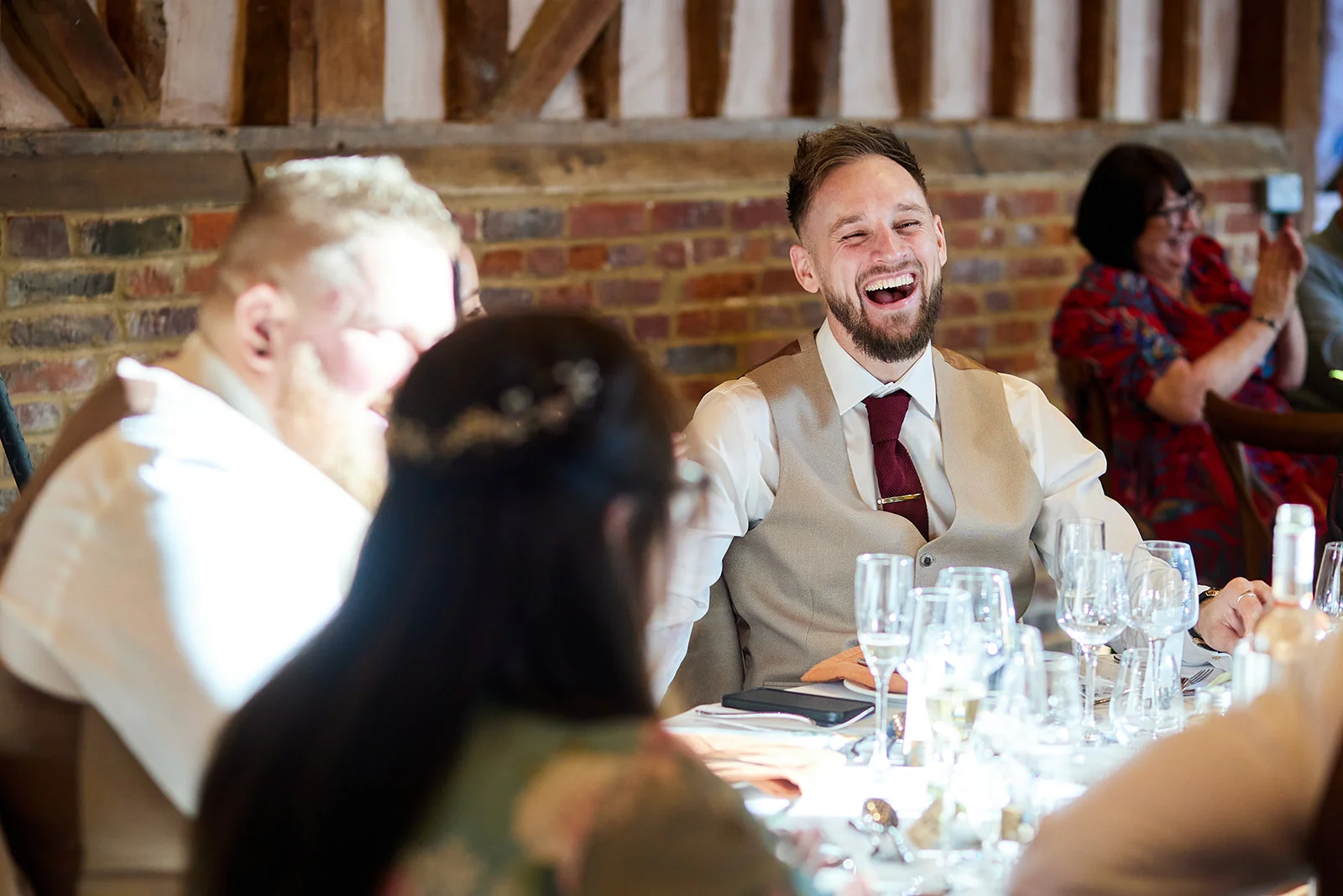A bearded groom in a beige waistcoat and red tie throws his head back laughing heartily during wedding speeches at a rustic barn reception.