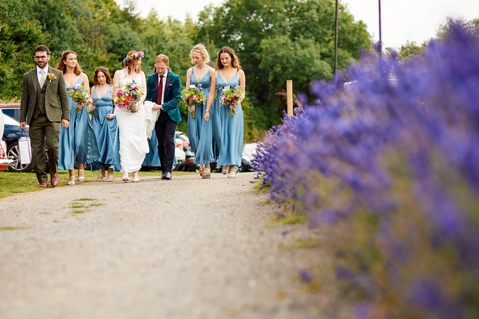 The bride, groom, and wedding party walk along an outdoor path lined with vibrant purple flowers during a summer wedding celebration.